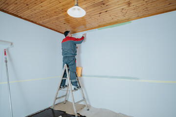 A man in a grey and red uniform paints a light blue wall standing on a ladder, completing a home...