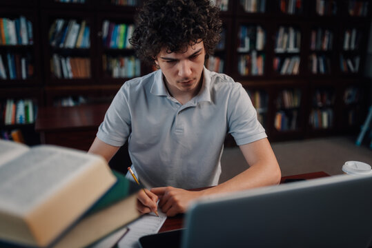 Focused student studying in library, writing notes and using laptop