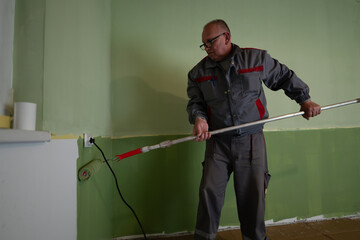 A professional worker paints a room using a green roller with an extension pole, creating a fresh...