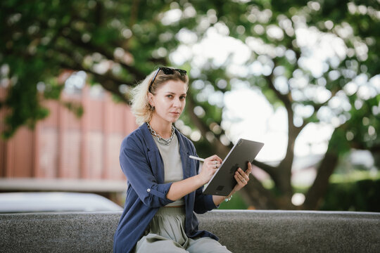 A confident woman uses a tablet and stylus outdoors, creating digital art with focus and style, embodying modern creativity, technology, and freelance lifestyle in an urban setting.