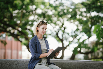 A confident woman uses a tablet and stylus outdoors, creating digital art with focus and style,...