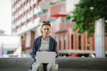 A cheerful young woman sits at a city park working with her laptop, combining work and leisure,...