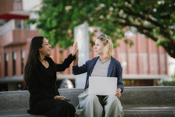 Two young women sit in a park discussing their work with their laptops, having a fun conversation,...