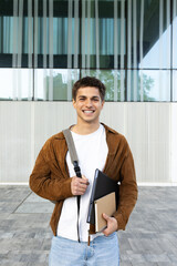 Smiling university student holding laptop and notebooks in front of modern building