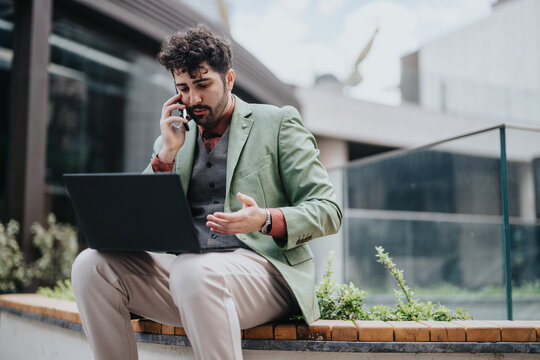 Confident young man multitasking by talking on a telephone and working on a laptop computer outside, showcasing remote work and modern connectivity.