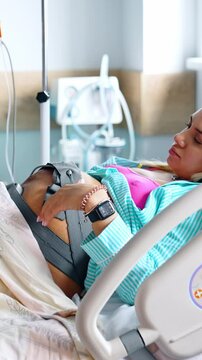 Blonde pregnant woman strokes her belly with attached cardiotocography belt. Future mom before childbirth in maternity hospital. Vertical video
