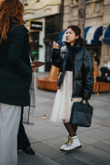 Two women are conversing in a lively urban street scene, showcasing unique outfits and stylish...