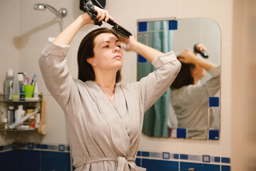 Woman in bathrobe drying her hair in the bathroom reflection. Self-care and beauty routine at home.