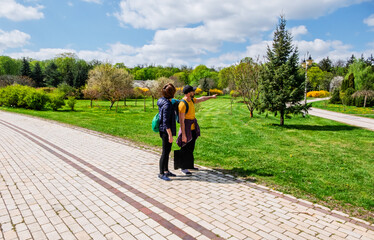 Two people stand on a paved path in a sunny park, looking and pointing towards something...