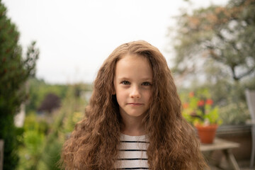 Close-up of sad girl with curly hair in striped shirt among potted flowers. Concept: childhood...