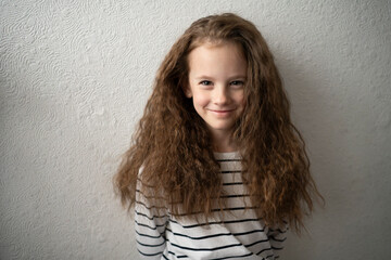 Little girl with long wavy hair smiling near a white wall indoors. Concept: everyday moments, carefree childhood, gentle energy, home atmosphere.