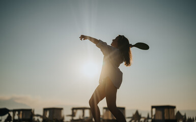 A woman joyfully plays paddle ball on the seashore at sunset, showing outdoor recreation.