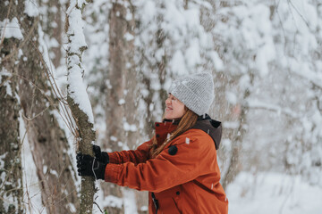 A cheerful woman dressed warmly stands in a snow-covered forest, admiring the serene nature while...