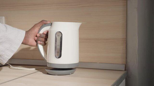Close-up of man in bathrobe holding electric kettle with hand over base, preparing to pour water. Sleek modern design of kettle and minimalist kitchen counter are highlighted in soft lighting