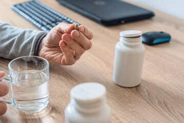 Closeup of man taking vitamin pills for flu prevention. Man holding supplement capsule at desk with water glass, bottles, keyboard, and laptop nearby