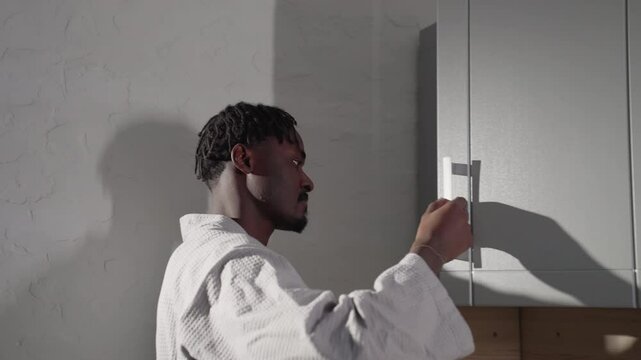 Man in bathrobe opening kitchen cabinet door in modern minimalist kitchen. Soft light highlights his profile and clean kitchen design, with shadows cast on the wall