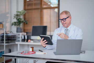 Senior doctor wearing lab coat and eyeglasses sitting at desk using tablet and laptop consulting patient online in clinic office, healthcare and medical technology concept