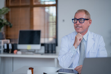 Thoughtful mature doctor wearing lab coat and glasses sitting at desk in bright medical office...