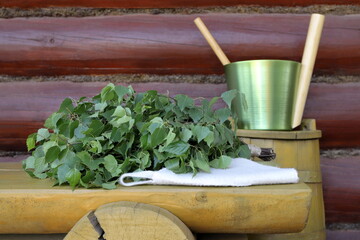 A birch broom is on the yellow wooden bench which is next to a yellow wooden barrel with a green metal sauna accessories on it, against a log wall.