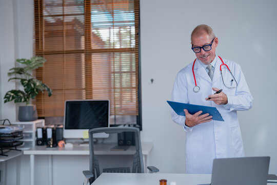 Smiling senior doctor wearing lab coat and stethoscope writing notes on clipboard while standing in medical office with computer and laptop