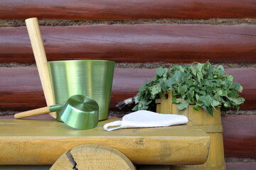 A green metal sauna accessories is on the yellow wooden bench which is next to a yellow wooden barrel with a birch broom on it, against a log wall.