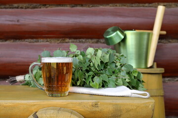 A glass mug of light beer and  birch broom are on the yellow wooden bench which is next to a yellow wooden barrel with a green metal sauna accessories on it, against a log wall.