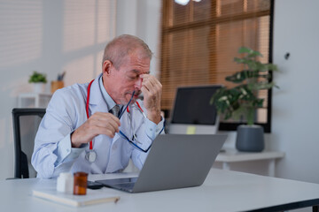 Senior doctor holding glasses and suffering from headache and stress while working with laptop in...