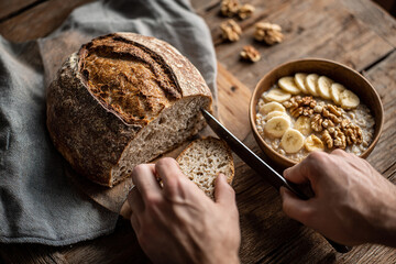 Close-up of hands slicing a fresh sourdough loaf beside a steaming bowl of oatmeal topped with sliced banana and walnuts, rustic wooden table with linen napkin