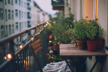 Tight view of a narrow city balcony with herb pots, folded blanket on chair, and string lights above, early morning