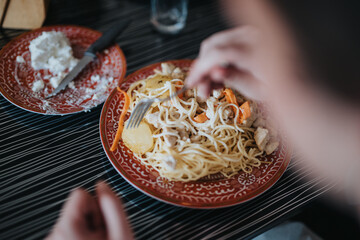 Close-up of a person eating a delicious pasta dish topped with chicken and vegetables on a decorative plate. The setting evokes a cozy dining experience.