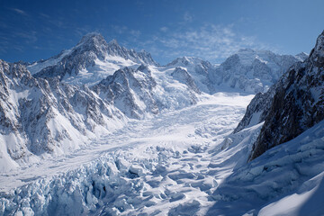 Wide shot of glacial surface with deep crevasses and distant snow-covered peaks, bright light