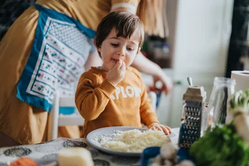 Fotobehang Kruidenier A child wearing a yellow shirt eats grated cheese from a plate while cooking alongside an adult in a bright and pleasant kitchen environment.  © qunica.com