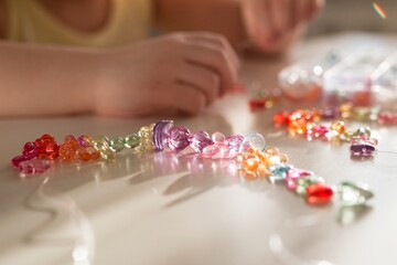 Hands of Child are Crafting Colorful Necklace or Bracelet from Beads in Sunlight on White Table.