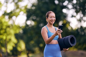 Smiling Woman in Activewear Holding Yoga Mat and Using Smartphone Outdoors