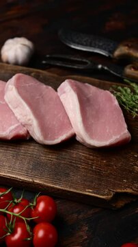 Raw pork escalopes arranged on rustic wooden board with fresh herbs and tomatoes