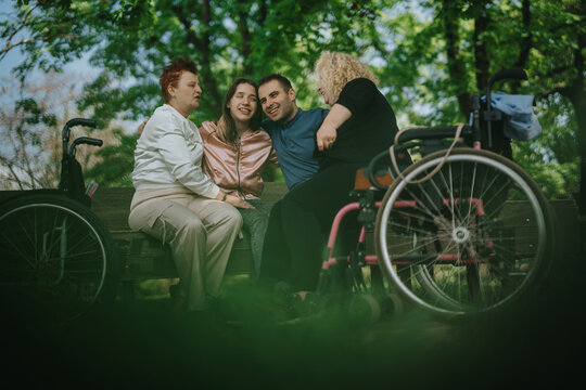 Group of friends with varying abilities spending quality time together seated on a bench outdoors, with wheelchairs nearby, fostering inclusion, friendship, and mutual joy.