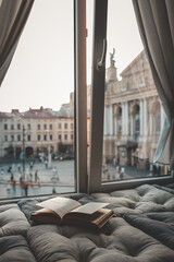 Cozy reading nook with an open book on a cushioned window seat overlooking a historic European city...