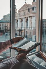 Person reading a Bible by the window with a view of the Lviv Opera House in the background during...