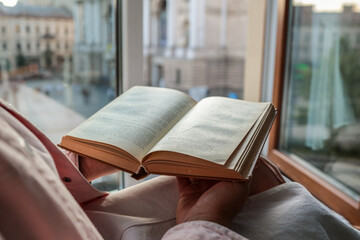 Person holding an open vintage book by a window with a view of a historic European square and opera...