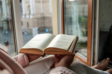 Person holding an open vintage book by a window with a view of a historic European square and opera...