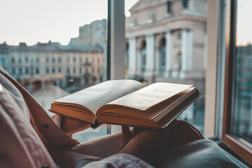 Person holding an open vintage book by a window with a view of a historic European square and opera...