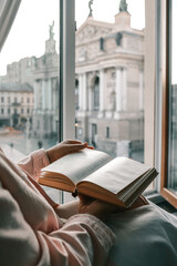 Person holding an open vintage book by a window with a view of a historic European square and opera...