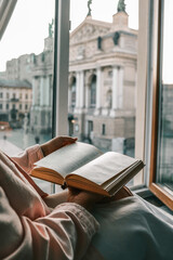 Person holding an open vintage book by a window with a view of a historic European square and opera...