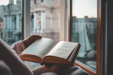 Person holding an open vintage book by a window with a view of a historic European square and opera...