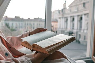 Person holding an open vintage book by a window with a view of a historic European square and opera...