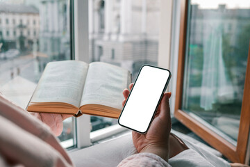Woman holding smartphone with blank screen while reading a book by the window with blurred view of Lviv Opera House in the background. Cozy, soft lighting and thoughtful atmosphere