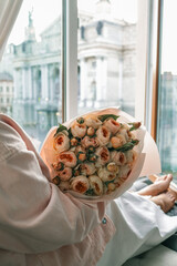 Woman holding a bouquet of delicate pastel roses by an open window with a view of Lviv Opera House....