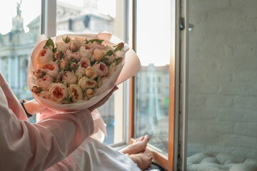 Woman holding a bouquet of delicate pastel roses by an open window with a view of Lviv Opera House....