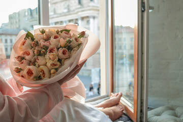 Woman holding a bouquet of delicate pastel roses by an open window with a view of Lviv Opera House....