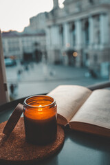 Close-up of a burning candle in an amber jar next to an open book on a window seat, with a soft...